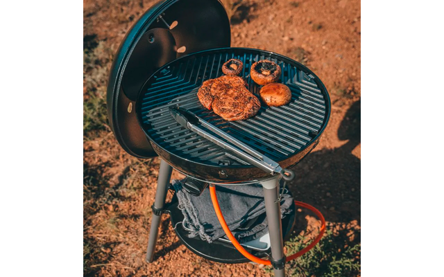 Runder Grill auf drei Beinen mit geöffnetem Deckel, Metallzange auf dem Rost, drei braune runde Stücke und zwei Pilze, orange Schlauch, trockener Boden