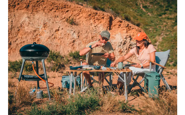 Zwei Personen sitzen an einem niedrigen Campingtisch vor einem felsigen Hang; links ein mobiler Grill; auf dem Tisch Geschirr, Besteck und Lebensmittel; beide tragen Kappen