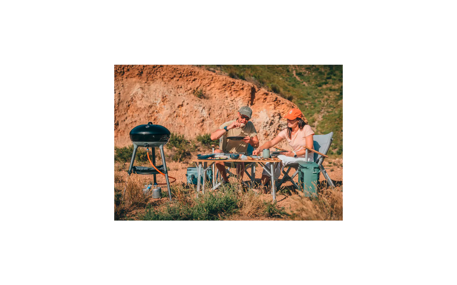 Zwei Personen sitzen an einem niedrigen Campingtisch vor einem felsigen Hang; links ein mobiler Grill; auf dem Tisch Geschirr, Besteck und Lebensmittel; beide tragen Kappen
