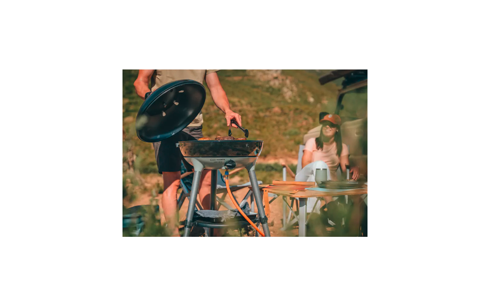 Person hebt den Deckel eines runden tragbaren Gasgrills und hält eine Grillzange; daneben ein Holztisch mit Tellern und Bechern; eine weitere Person sitzt am Tisch mit Mütze und Sonnenbrille; grasbewachsene Hügel im Hintergrund