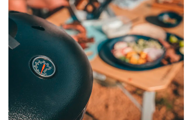Schwarzer Grilldeckel mit eingebautem Thermometer im Vordergrund, unscharfe Teller mit Salat und Besteck auf einem Holztisch im Hintergrund