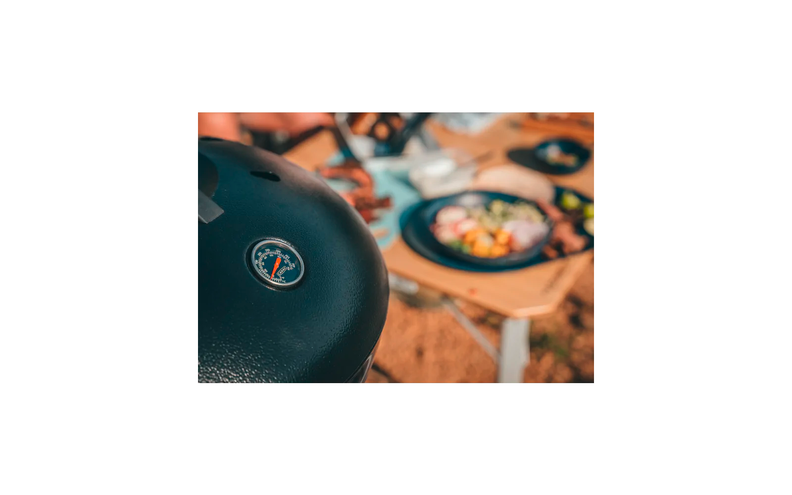 Schwarzer Grilldeckel mit eingebautem Thermometer im Vordergrund, unscharfe Teller mit Salat und Besteck auf einem Holztisch im Hintergrund