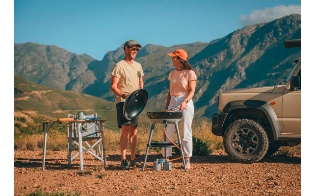 Zwei Personen stehen auf Schotter neben einem Geländewagen; eine Person hält eine runde Pfanne, die andere steht neben einem tragbaren Grill; Campingtisch mit Utensilien und Berglandschaft im Hintergrund