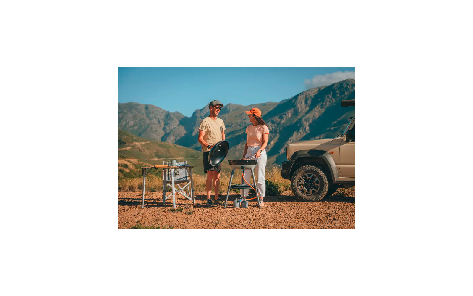 Zwei Personen stehen auf Schotter neben einem Geländewagen; eine Person hält eine runde Pfanne, die andere steht neben einem tragbaren Grill; Campingtisch mit Utensilien und Berglandschaft im Hintergrund