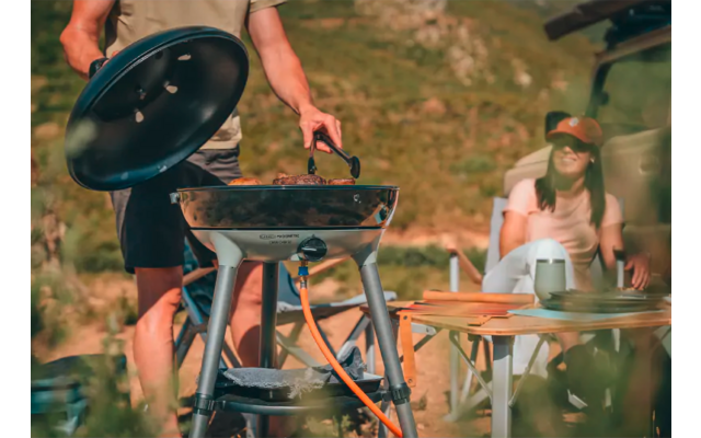 Person hebt den Deckel eines runden tragbaren Gasgrills und hält eine Grillzange; daneben ein Holztisch mit Tellern und Bechern; eine weitere Person sitzt am Tisch mit Mütze und Sonnenbrille; grasbewachsene Hügel im Hintergrund