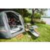 Geöffnetes Vorzelt an einem See; eine Person sitzt im Zelt an einem Tisch mit Laptop und Campingausrüstung, eine weitere Person liegt auf einer Liege auf der Wiese; Fußmatte mit «welcome», Sandalen, Klapptisch, Bäume und See im Hintergrund
