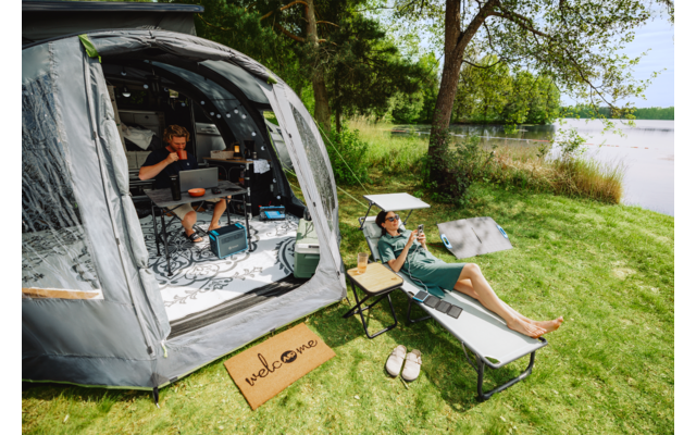 Geöffnetes Vorzelt an einem See; eine Person sitzt im Zelt an einem Tisch mit Laptop und Campingausrüstung, eine weitere Person liegt auf einer Liege auf der Wiese; Fußmatte mit «welcome», Sandalen, Klapptisch, Bäume und See im Hintergrund