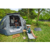 Offenes Campingzelt auf einer Wiese; eine Person sitzt im Zelt mit Laptop, eine Person liegt auf einer Liege und hält ein Fernglas; kleiner Beistelltisch, Hausschuhe, Fußmatte mit Schriftzug, Solarpanel auf dem Gras, Bäume und See im Hintergrund