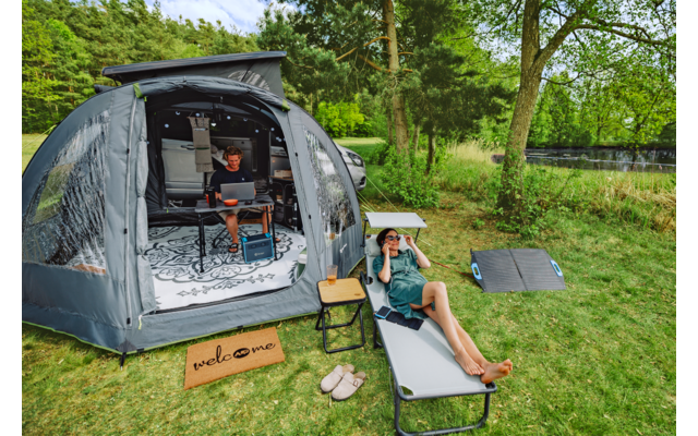 Offenes Campingzelt auf einer Wiese; eine Person sitzt im Zelt mit Laptop, eine Person liegt auf einer Liege und hält ein Fernglas; kleiner Beistelltisch, Hausschuhe, Fußmatte mit Schriftzug, Solarpanel auf dem Gras, Bäume und See im Hintergrund