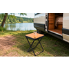 Kleiner Klapptisch mit Glas Wasser neben offenem Campingvan, sichtbares Bett im Innenraum, Grasboden und See mit Baumreihe im Hintergrund