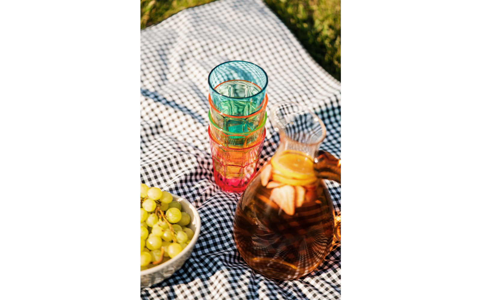 Mehrere gestapelte bunte Gläser, eine Karaffe mit orangefarbener Flüssigkeit und Zitronenscheiben, eine Schale mit grünen Trauben auf einer schwarz-weiß karierten Decke im Gras