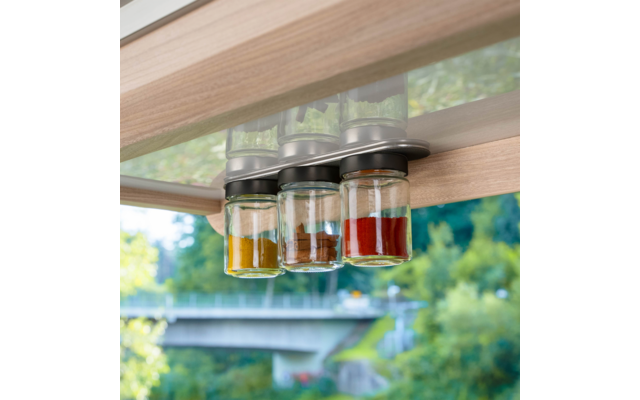 Drei Gläser mit Metalldeckeln hängen unter einem Holzregal; sie enthalten gelbes, braunes und rotes Pulver. Im Hintergrund sind Bäume und eine verschwommene Brücke durch ein Fenster sichtbar