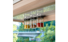 Drei Gläser mit Metalldeckeln hängen unter einem Holzregal; sie enthalten gelbes, braunes und rotes Pulver. Im Hintergrund sind Bäume und eine verschwommene Brücke durch ein Fenster sichtbar
