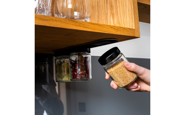 Hand hält ein kleines Glas mit schwarzem Deckel und gelben Körnern unter der Kante eines hölzernen Küchenschranks; zwei weitere Glasgefäße sind befestigt