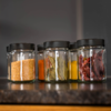 Fünf kleine Glasgläser mit schwarzen Deckeln nebeneinander auf einer Arbeitsfläche; enthalten gelbe Senfkörner, orange‑rotes Pulver, Lorbeerblätter, gelbes Pulver und getrocknete rote Chilischoten