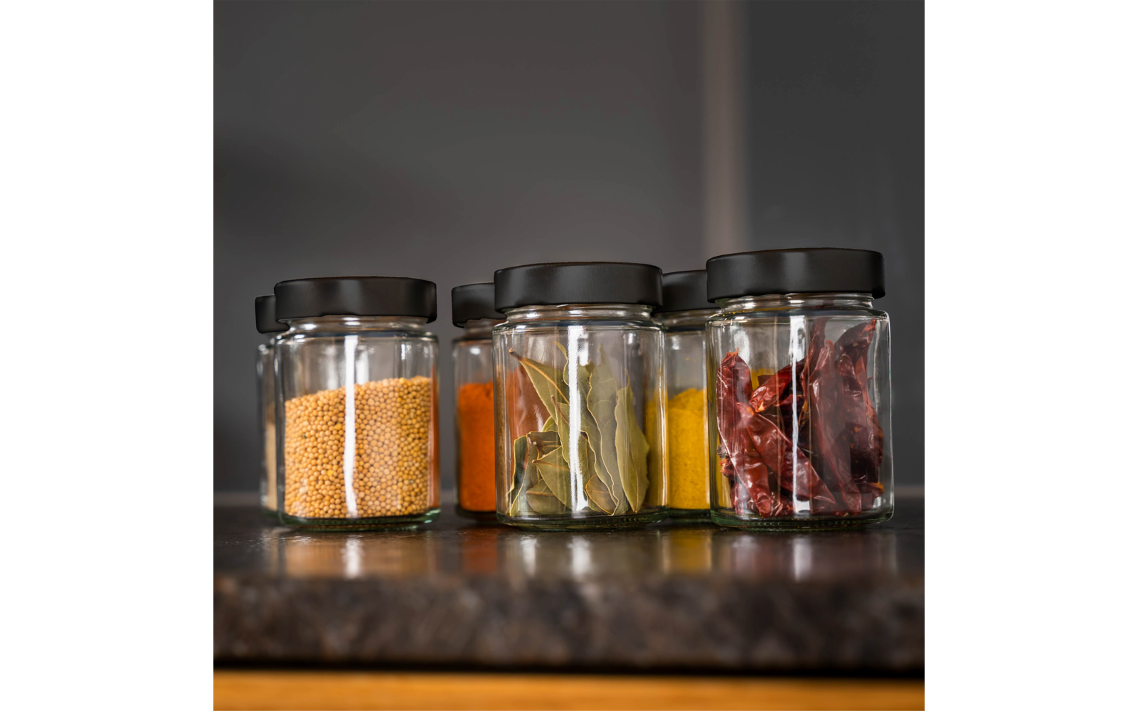 Fünf kleine Glasgläser mit schwarzen Deckeln nebeneinander auf einer Arbeitsfläche; enthalten gelbe Senfkörner, orange‑rotes Pulver, Lorbeerblätter, gelbes Pulver und getrocknete rote Chilischoten