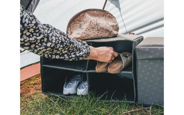Mano che mette un sandalo marrone in uno scomparto di uno scaffale aperto; sneaker bianche a sinistra, pochette con motivo sopra, cubo grigio a destra, erba in primo piano, tessuto della tenda sullo sfondo