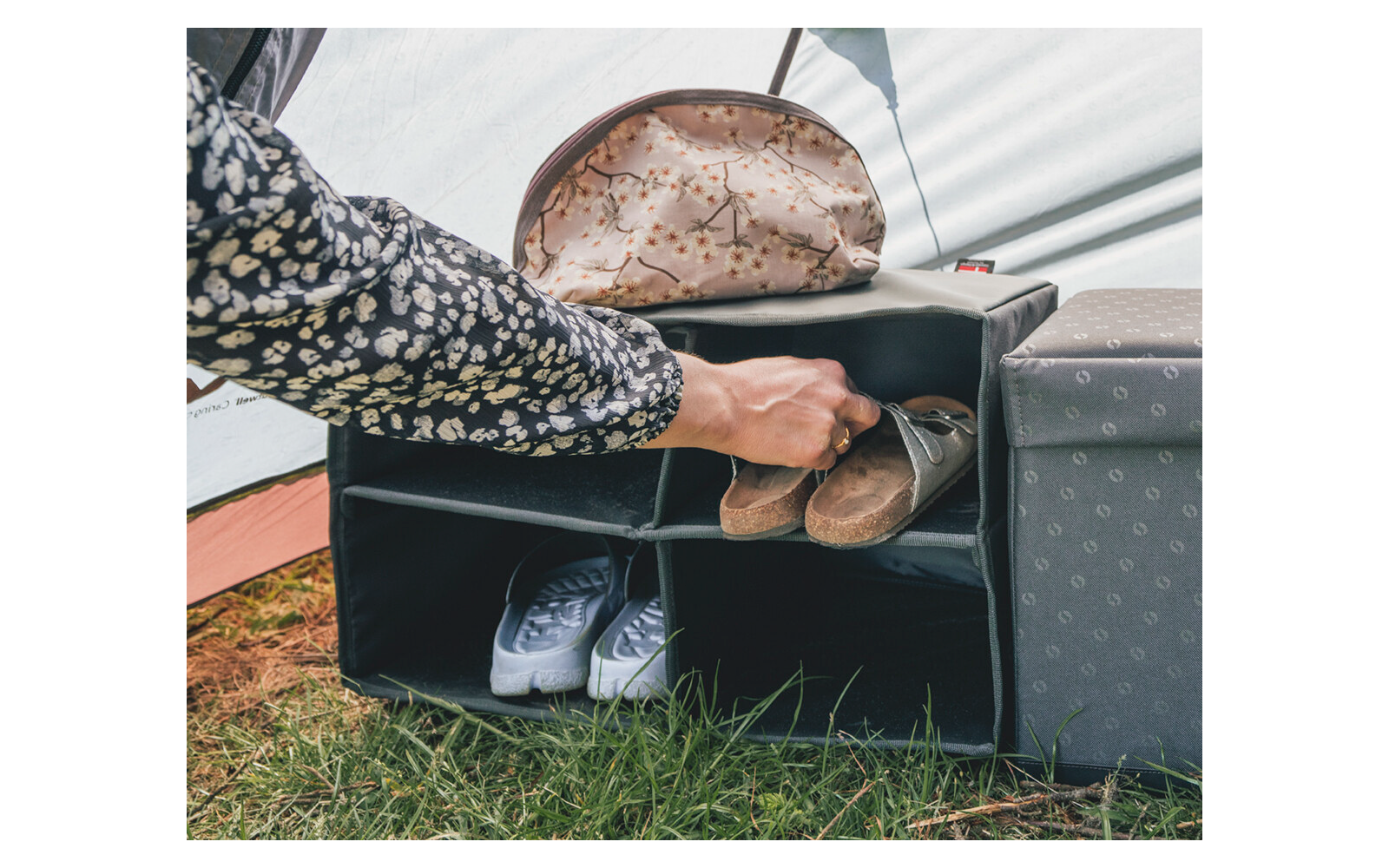 Mano che mette un sandalo marrone in uno scomparto di uno scaffale aperto; sneaker bianche a sinistra, pochette con motivo sopra, cubo grigio a destra, erba in primo piano, tessuto della tenda sullo sfondo