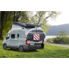 Camper bianco con tetto rialzabile aperto e borsa posteriore con pannello di segnalazione rosso-bianco, parcheggiato su prato vicino a un lago; alberi e colline sullo sfondo
