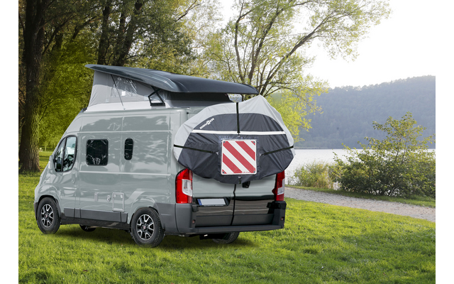 Camper bianco con tetto rialzabile aperto e borsa posteriore con pannello di segnalazione rosso-bianco, parcheggiato su prato vicino a un lago; alberi e colline sullo sfondo