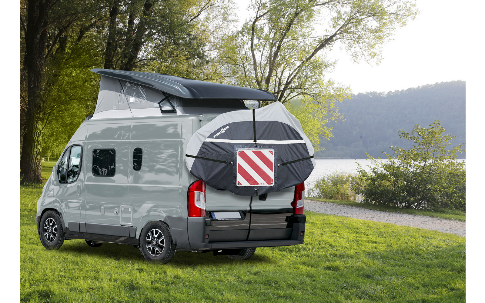 Camper bianco con tetto rialzabile aperto e borsa posteriore con pannello di segnalazione rosso-bianco, parcheggiato su prato vicino a un lago; alberi e colline sullo sfondo
