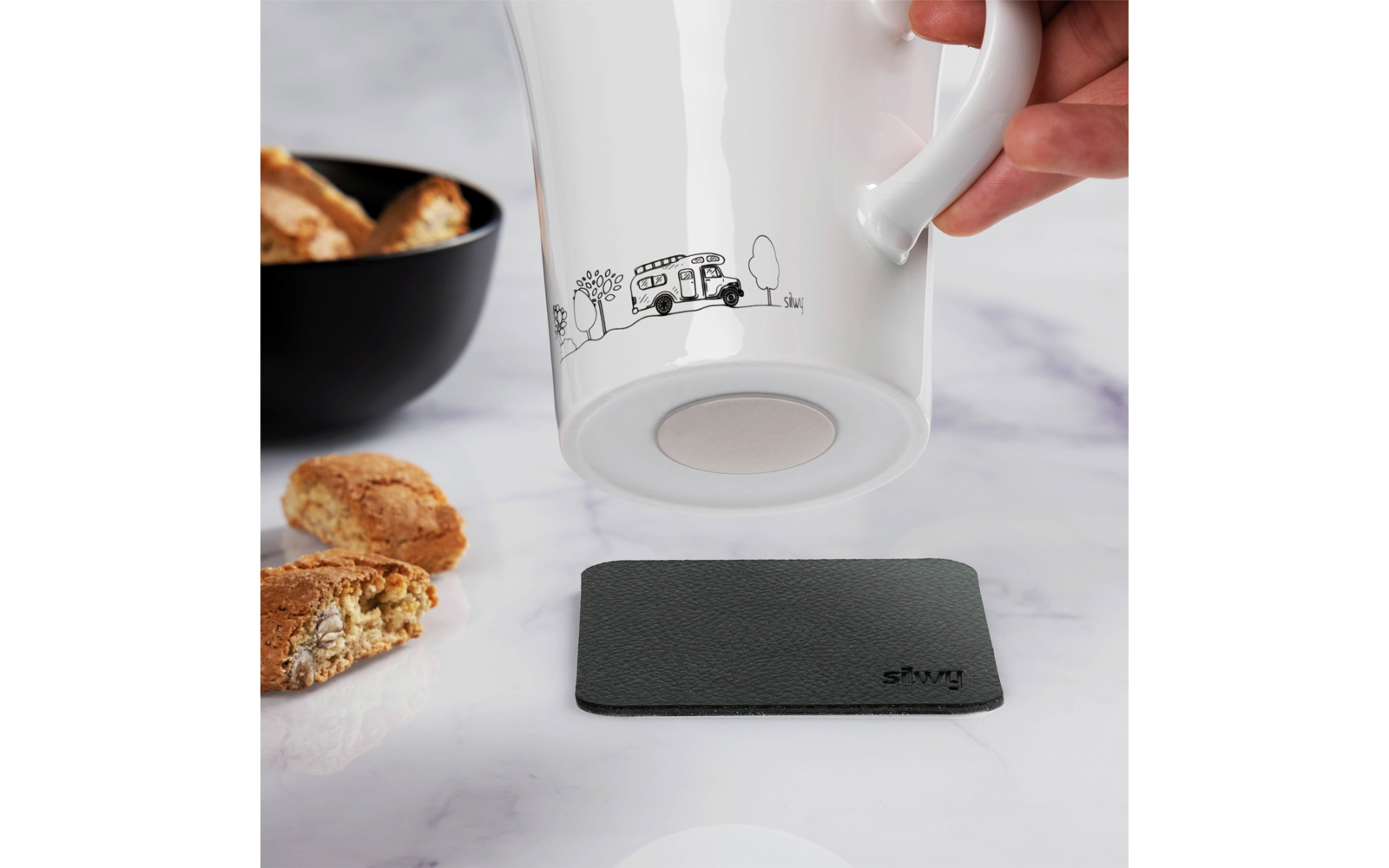 Hand hält eine weiße Tasse mit einem gezeichneten Wohnmobil, darüber ein schwarzer Untersetzer auf einem Marmortisch, daneben Kekse und eine schwarze Schüssel
