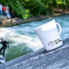 Weiße Tasse mit schwarzem Muster steht auf steinerner Fläche, im Hintergrund Fluß mit Surfer und Bäumen