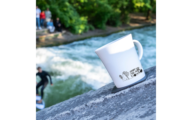 Weiße Tasse mit schwarzem Muster steht auf steinerner Fläche, im Hintergrund Fluß mit Surfer und Bäumen
