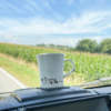 Weiße Tasse mit schwarzem Aufdruck steht auf dem Armaturenbrett eines Fahrzeugs, im Hintergrund Felder und blauer Himmel