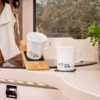 Zwei weiße Tassen mit schwarzem Muster stehen auf einer Küchenarbeitsplatte, eine Tasse lehnt auf einem Holzuntersetzer
