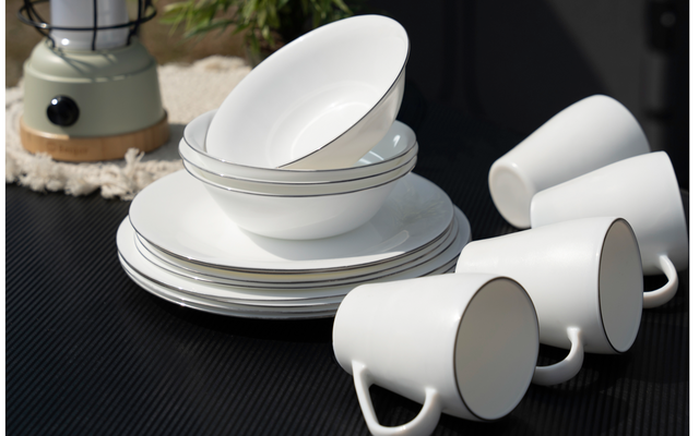 Stacked white plates and bowls beside three white cups on a dark surface, with a lantern and plant in the background