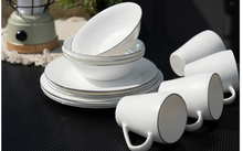 Stacked white plates and bowls beside three white cups on a dark surface, with a lantern and plant in the background