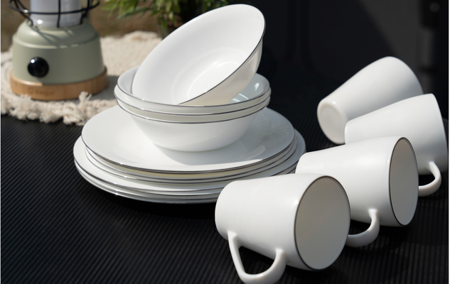 Stacked white plates and bowls beside three white cups on a dark surface, with a lantern and plant in the background