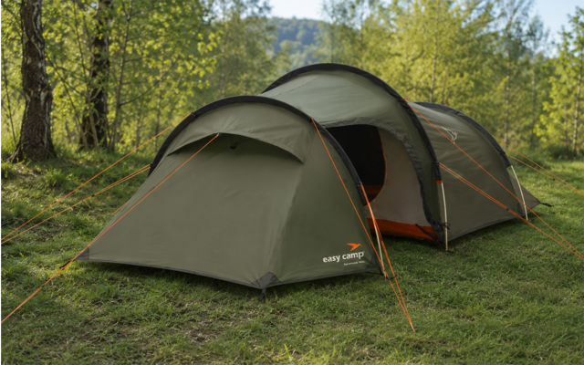 Grünes Campingszelt mit zwei verbundenen Sektionen auf Gras, orangefarbene Abspannleinen, Bäume und Hügel im Hintergrund