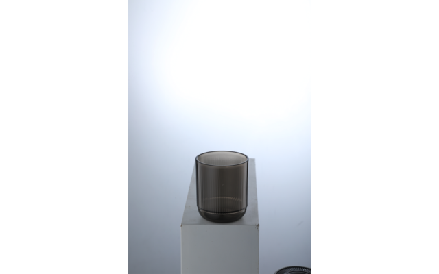 Verre foncé côtelé posé sur un socle rectangulaire blanc devant un fond clair en dégradé ; en bas à droite, une forme ronde sombre partiellement visible