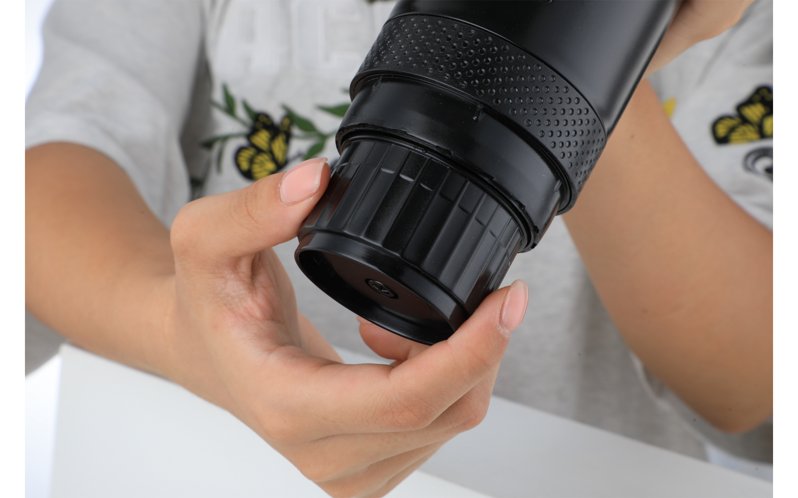 Deux mains tiennent un objectif d'appareil photo noir ; chemise blanche visible avec broderies florales jaunes et surface de table blanche
