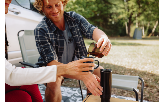 Person gießt Kaffee aus einer Kanne in eine Tasse, eine zweite Person hält die Tasse; Thermoskanne auf kleinem Tisch, Klappstühle, Auto und Bäume auf einer Wiese im Hintergrund