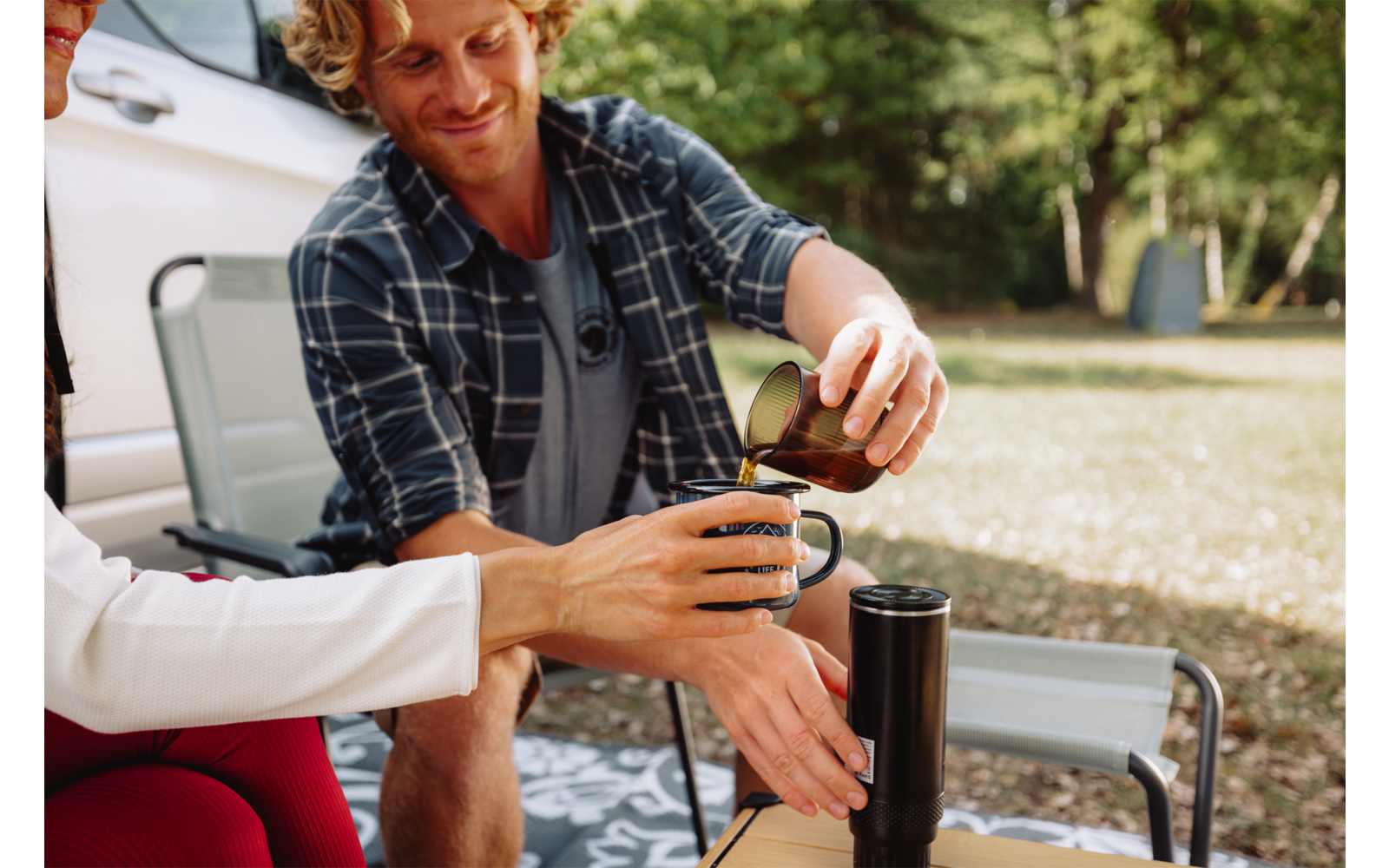 Person gießt Kaffee aus einer Kanne in eine Tasse, eine zweite Person hält die Tasse; Thermoskanne auf kleinem Tisch, Klappstühle, Auto und Bäume auf einer Wiese im Hintergrund