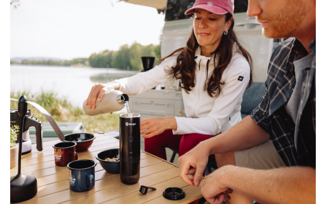 Frau gießt Flüssigkeit in eine tragbare Kaffeemaschine auf einem Campingtisch; Mann sitzt daneben; Tassen und Zubehör auf dem Tisch; im Hintergrund Campingbus und ein See mit Bäumen