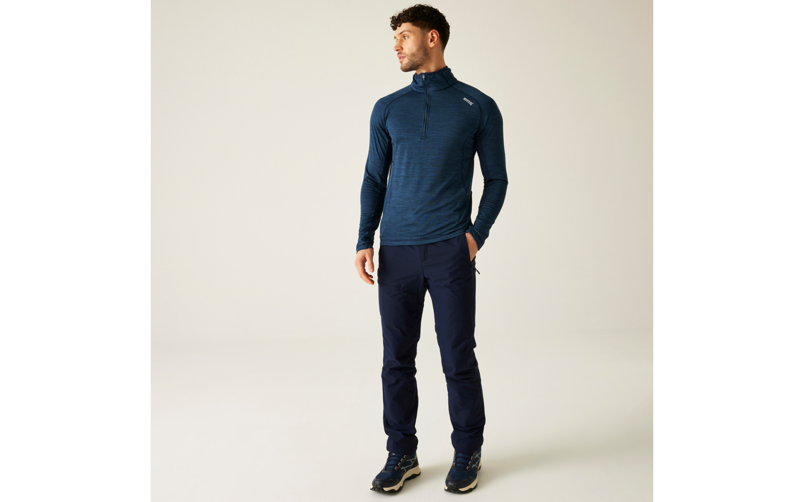 Ein Mann steht vor einem hellen Hintergrund, trägt ein dunkelblaues Langarmshirt und dunkelblaue Hosen