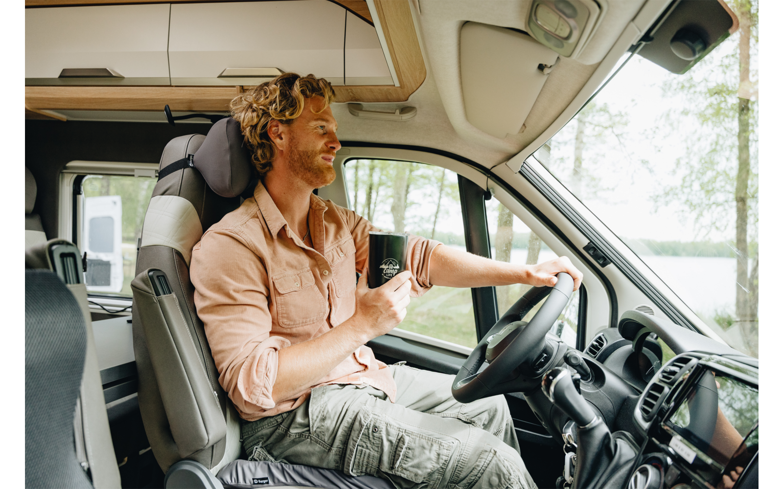Adulto con capelli ricci seduto sul sedile del conducente di un camper, tiene una tazza; volante, cruscotto e alberi e un lago visibili dalla finestra