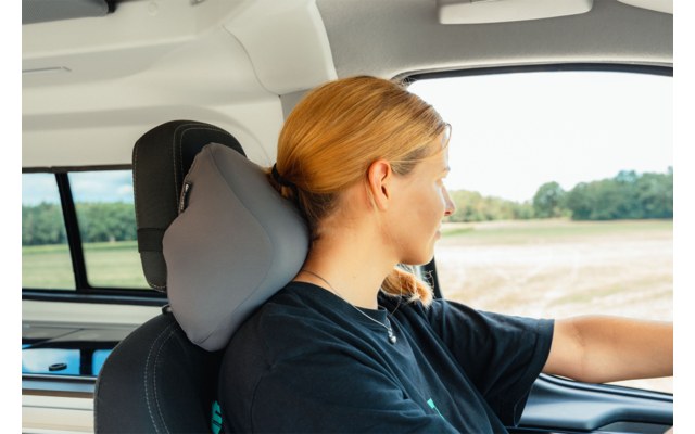 Woman with tied-back blonde hair seated at the wheel, head resting on a gray travel pillow, looking right, car window showing fields outside