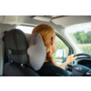 Rear view of a person with braided blonde hair at the wheel, gray neck pillow attached to the headrest, car interior, green landscape visible through the window