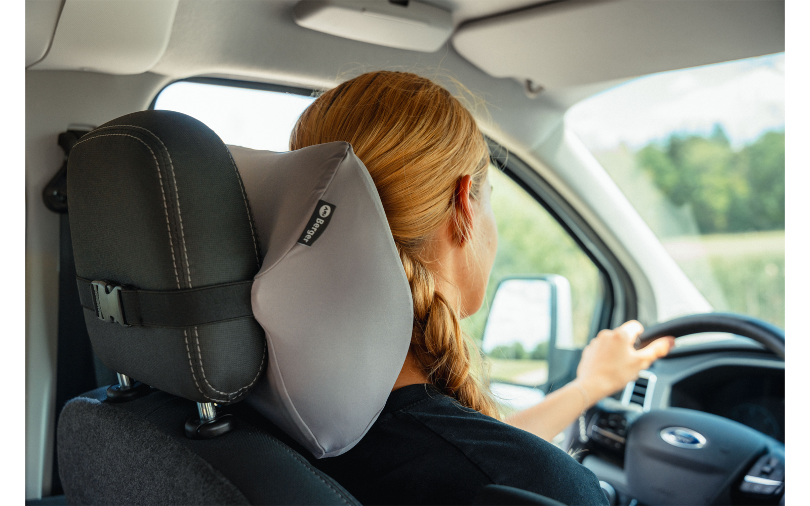 Rear view of a person with braided blonde hair at the wheel, gray neck pillow attached to the headrest, car interior, green landscape visible through the window