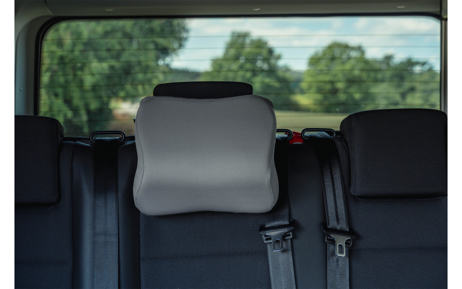 Car rear seat with a gray pad on the center headrest, black seats, visible seat belts and rear window showing trees