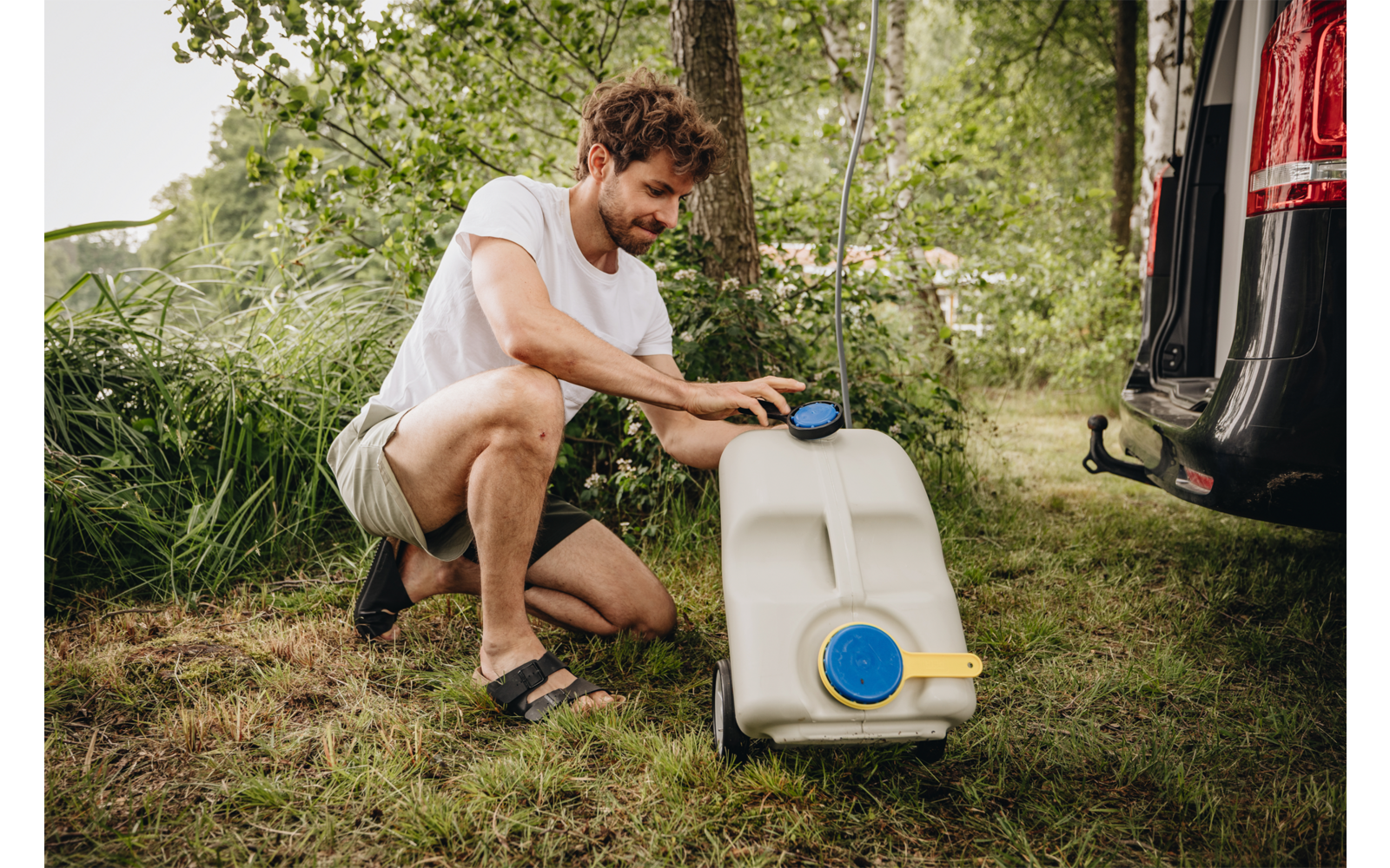 A person kneeling on grass next to a white water container with a blue cap and yellow handle, beside a parked van