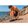 Hombre agachado en una playa rocosa poniéndose zapatos de agua azules, mar y rocas al fondo