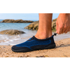 Primer plano de un pie con un zapato de agua azul en una playa de arena; una mano aprieta el cordón en el talón, rocas y mar al fondo
