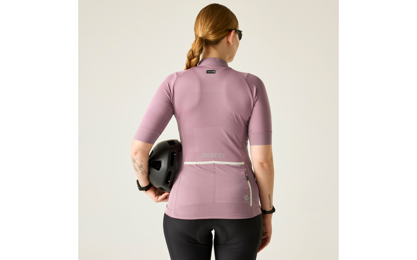 Rückenansicht einer Person in rosafarbenem Radtrikot und schwarzer Radhose, geflochtener Zopf, Sonnenbrille, hält einen schwarzen Fahrradhelm in der rechten Hand; heller Hintergrund