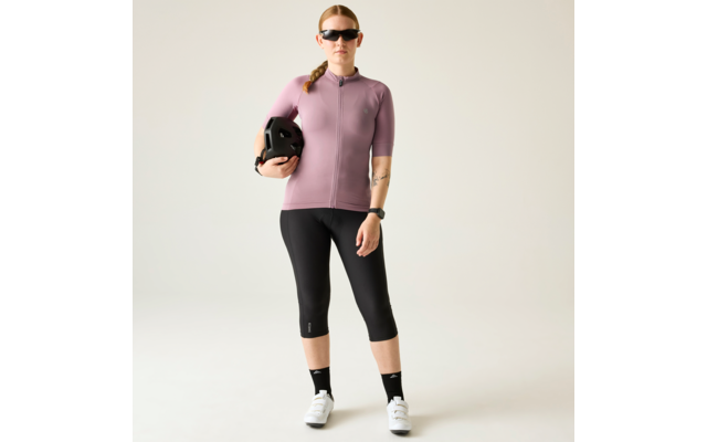 Person steht vor neutralem Hintergrund, trägt kurzärmeliges rosafarbenes Oberteil, schwarze knielange Sporthose, Sonnenbrille, hält schwarzen Helm in der linken Armbeuge, geflochtener Zopf, schwarze Knöchelsocken, weiße Sneaker, Uhr am rechten Handge